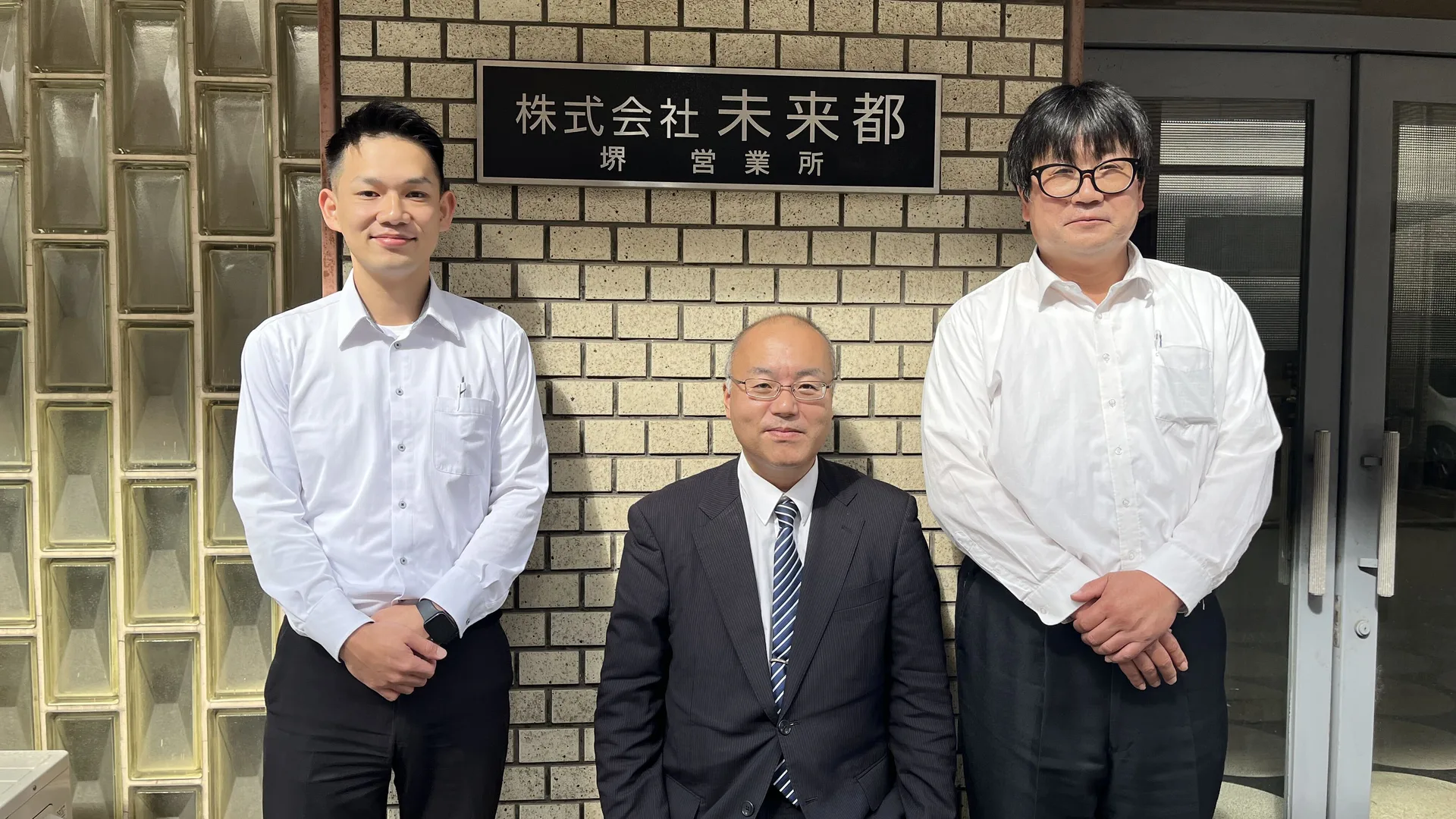 堺営業所の看板の前で、同僚と並ぶS.M.さん